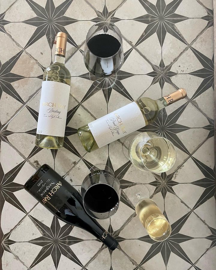 Three bottles of Arch Ray wine lay flat on their backs atop a tile floor. The tile is off-white with a pattern of geometric grey stars creating a lattice effect. Mixed amongst the bottles, there are four different wine glasses, each filled with a different wine. 