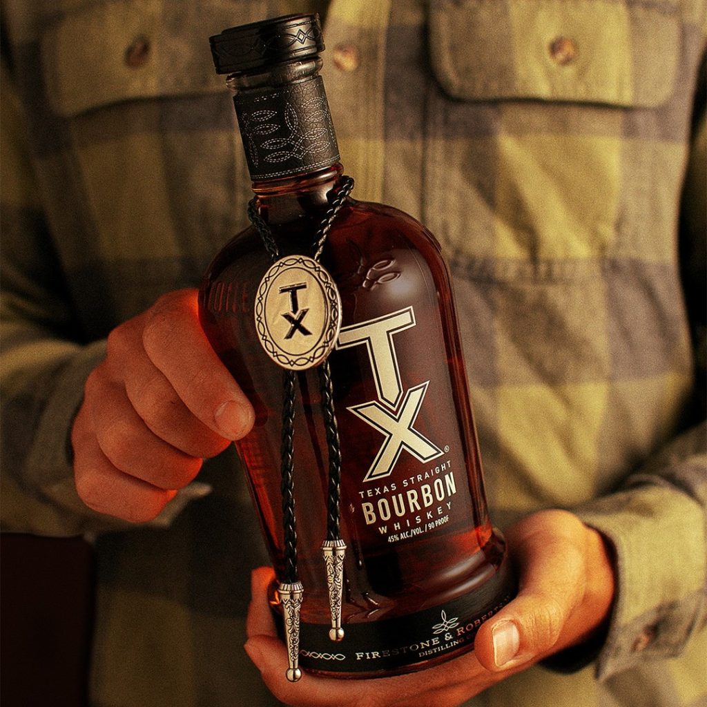 Close up shot of a man holding a bottle of TX Whiskey with a distinctive leather neckwrap, as well as a lather bolero tie with the TX Whiskey logo wrapped around the neck of the bottle. The man wears a green and brown plaid shirt. 