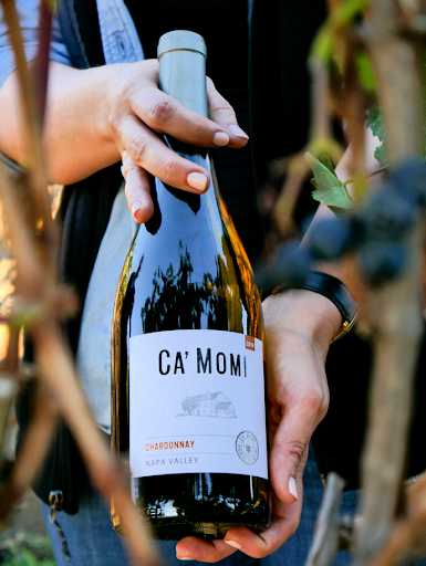 Close up shot of a woman's hands holding a bottle of Ca' Momi Chardonnay. In the foreground, blurry grape vines creep in at the edges to frame the bottle. 