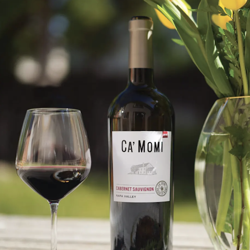 A bottle of Ca' Momi Cabernet Sauvignon sits on a white table next to a glass of the red wine, as well as a clear vase filled with yellow tulips. 