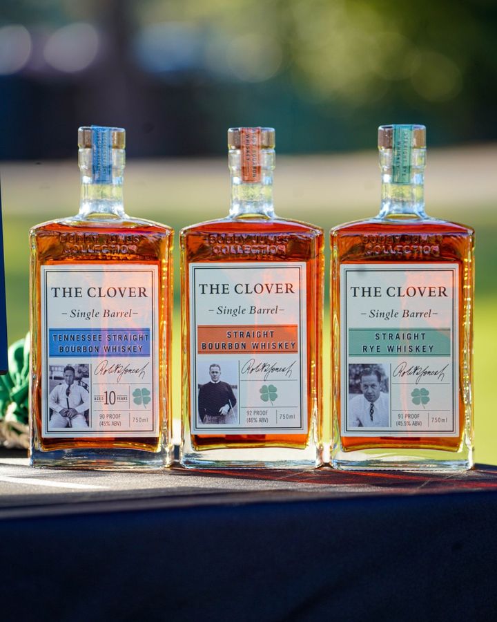 Three Clover Whiskey bottles sitting on a dark wood table outside, the background is blurred. 