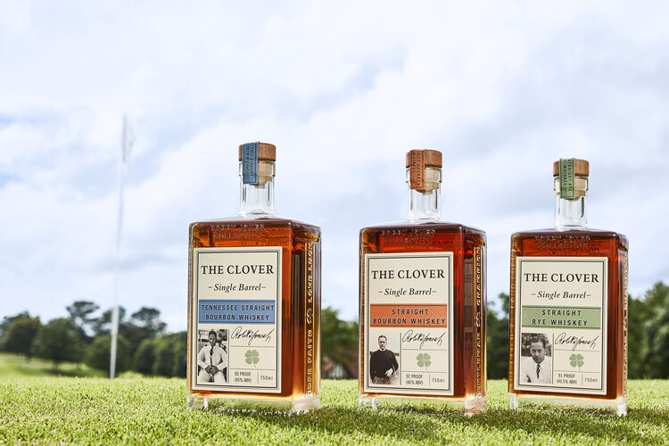 Three Clover Whiskey glass bottles sitting on an outside putting green with a golf flag and blue sky in the background. 
