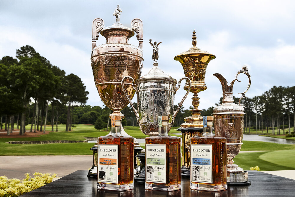 Three Clover Whiskey glass bottles sitting on a dark wood table with four golf championship trophies behind them, there is a golf course in the background. 