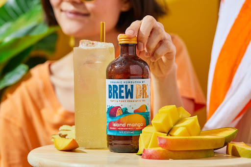 Bottle of Brew Dr Kombucha next to fresh cut mango and a glass of a yellow beverage. In the background, we see a woman in an orange shirt, whose hands it reaching for the cap of the kombucha bottle.