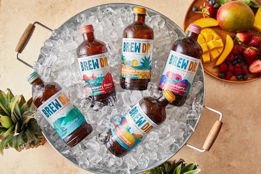 Top-down view of five bottles of Brew Dr kombucha laying in a metal tub full of ice, with freshly cut fruit in the background.