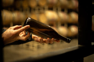 An empty Revino refillable bottle seen in someone's hands as they inspect markings on the bottom; unfocused wine barrels line the wall in the background.